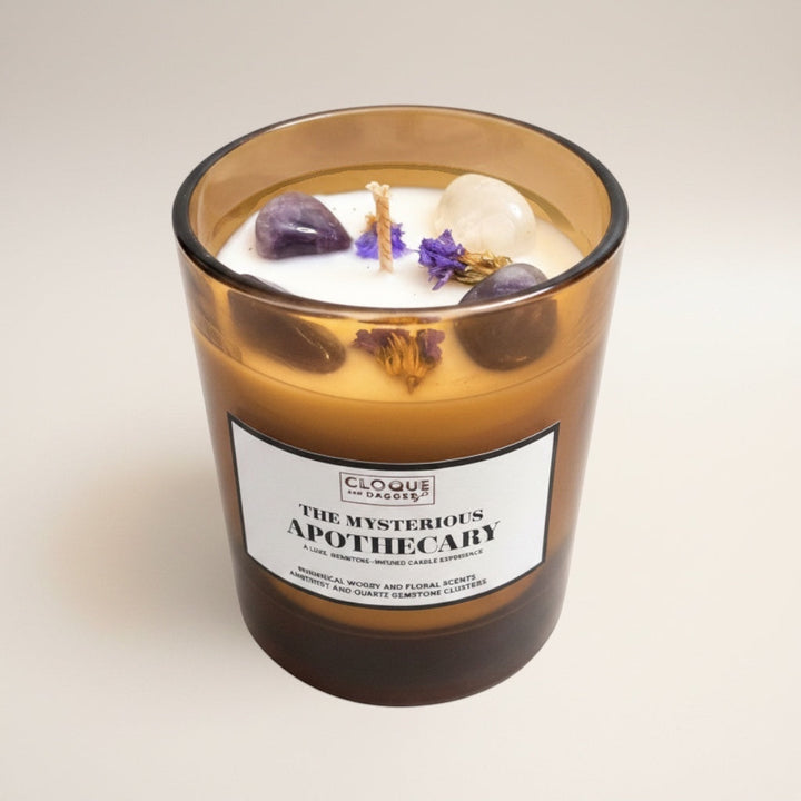Cloque and Dagger Mysterious Apothecary candle in amber glass with amethyst, clear quartz, and botanicals. Whimsical, woody, floral scents.